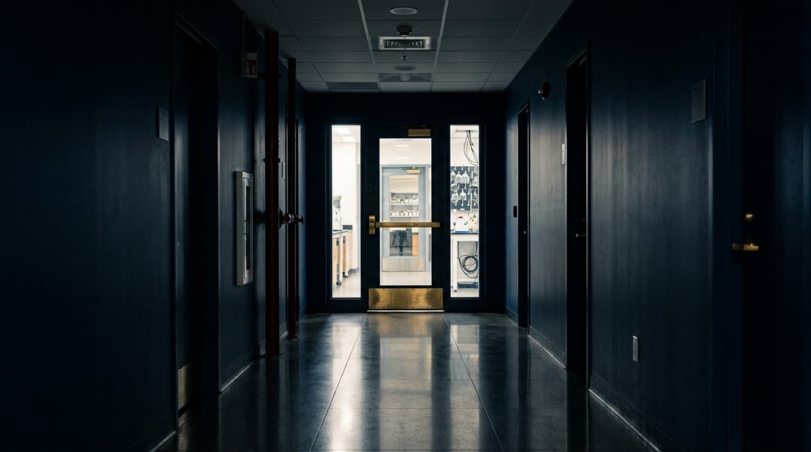 Corridoio di laboratorio in penombra con porta a vetri illuminata in fondo.